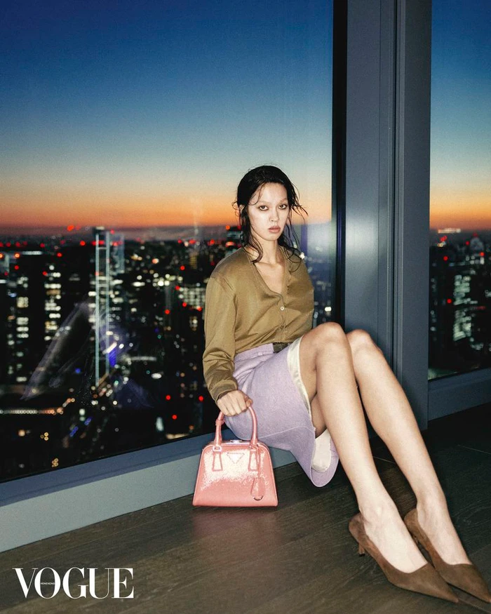 Model seated on a high-rise floor-to-ceiling window ledge holding a pink handbag with a city skyline at sunset in the background