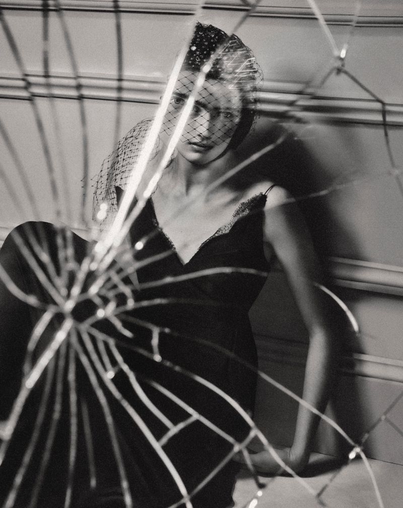 Monochrome portrait of a woman in a dark dress and veil, reflected in a shattered mirror, in a studio with wall paneling.