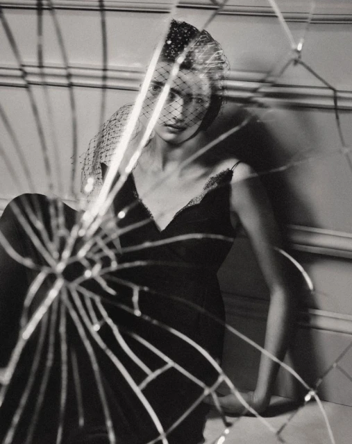 Monochrome portrait of a woman in a dark dress and veil, reflected in a shattered mirror, in a studio with wall paneling.