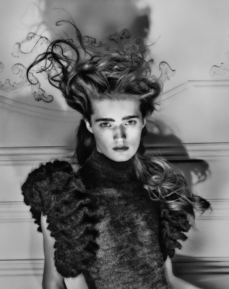 Black and white portrait of a woman with stylized hair and a dark, textured top in front of an ornate wall, showcasing high-fashion elegance.
