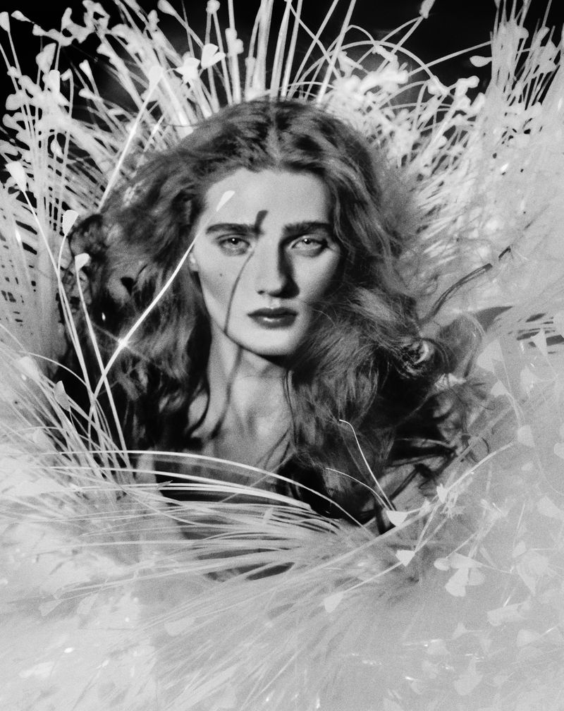 Black and white portrait of a woman with elaborate feathered hair and a serious expression, captured in a studio setting with dramatic lighting.