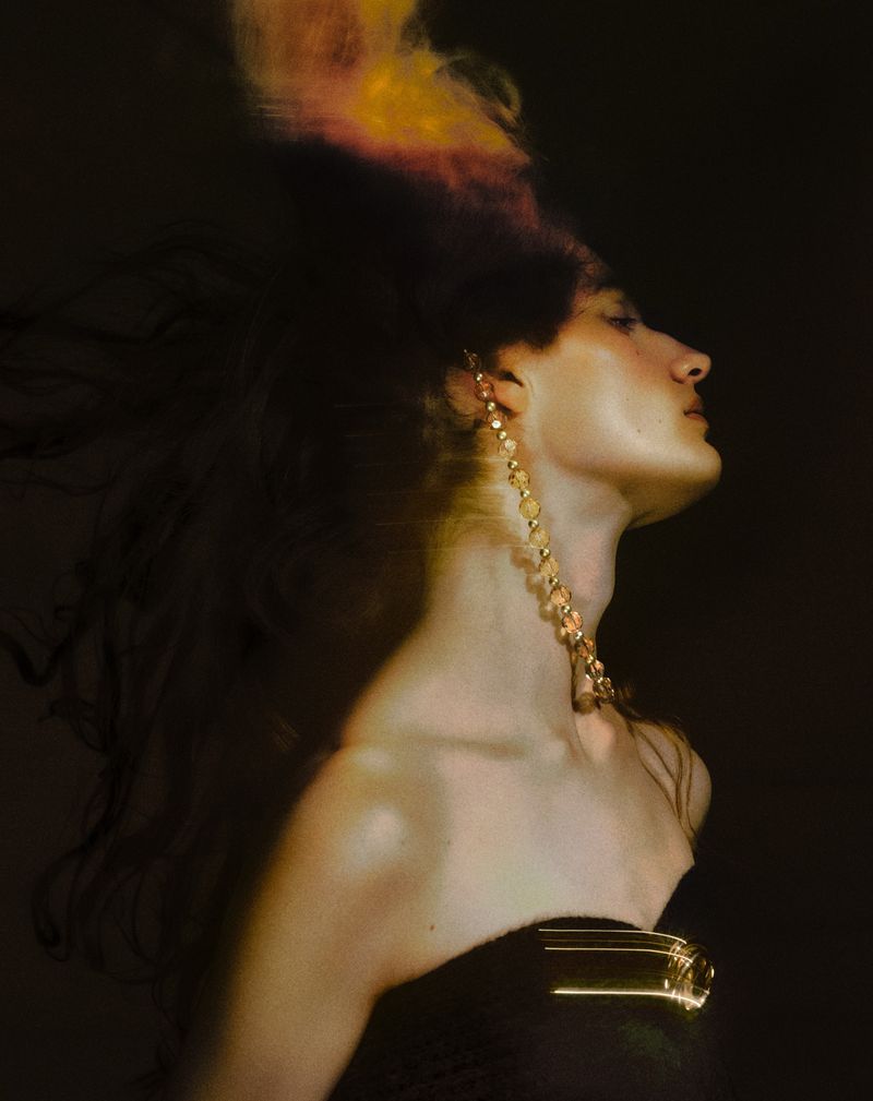 Elegant woman in profile wearing gold earrings and a strapless dress, with blown-back hair, against a dark, moody background.