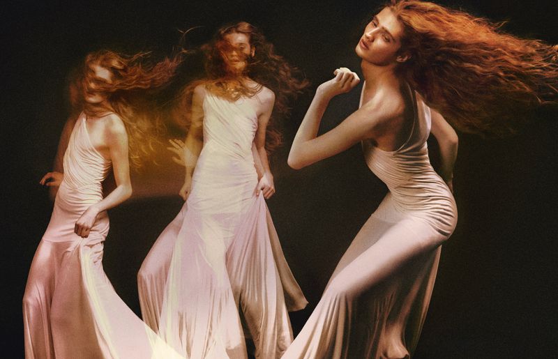 Three red-haired models in long white dresses pose in a dark photo studio with slow-shutter motion blur, creating a dreamlike fashion image.