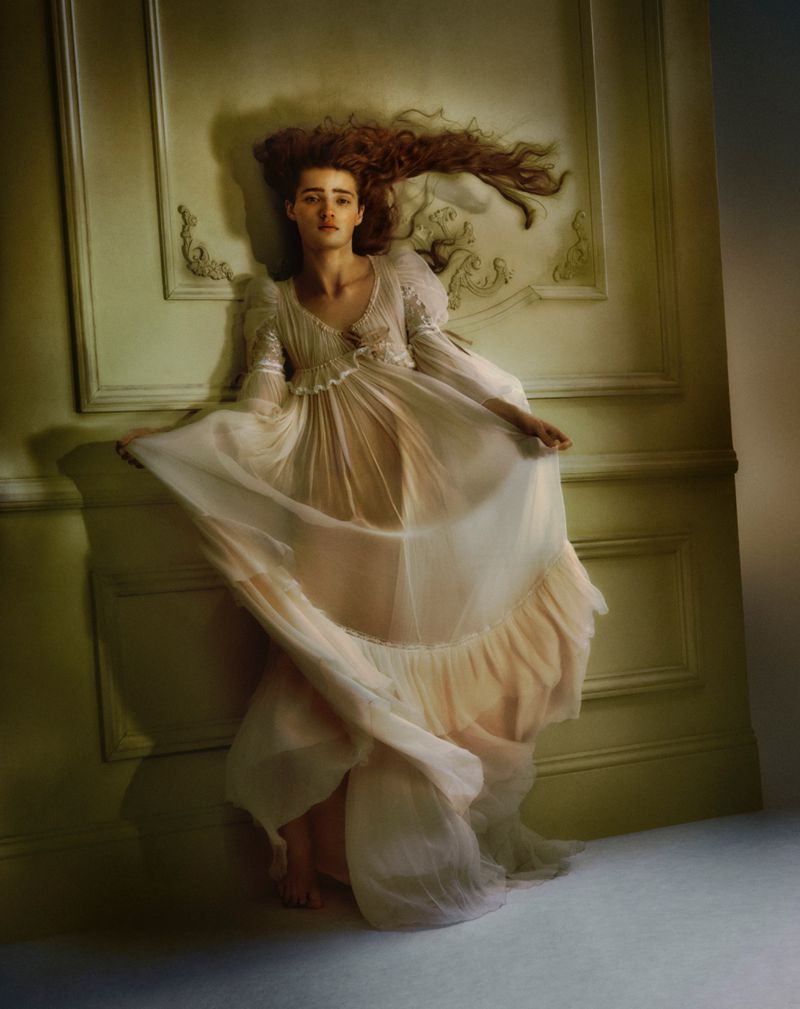 A fair-skinned woman with long red hair lies against a pale green wall wearing a sheer, flowing white dress in a soft-focus studio shot.