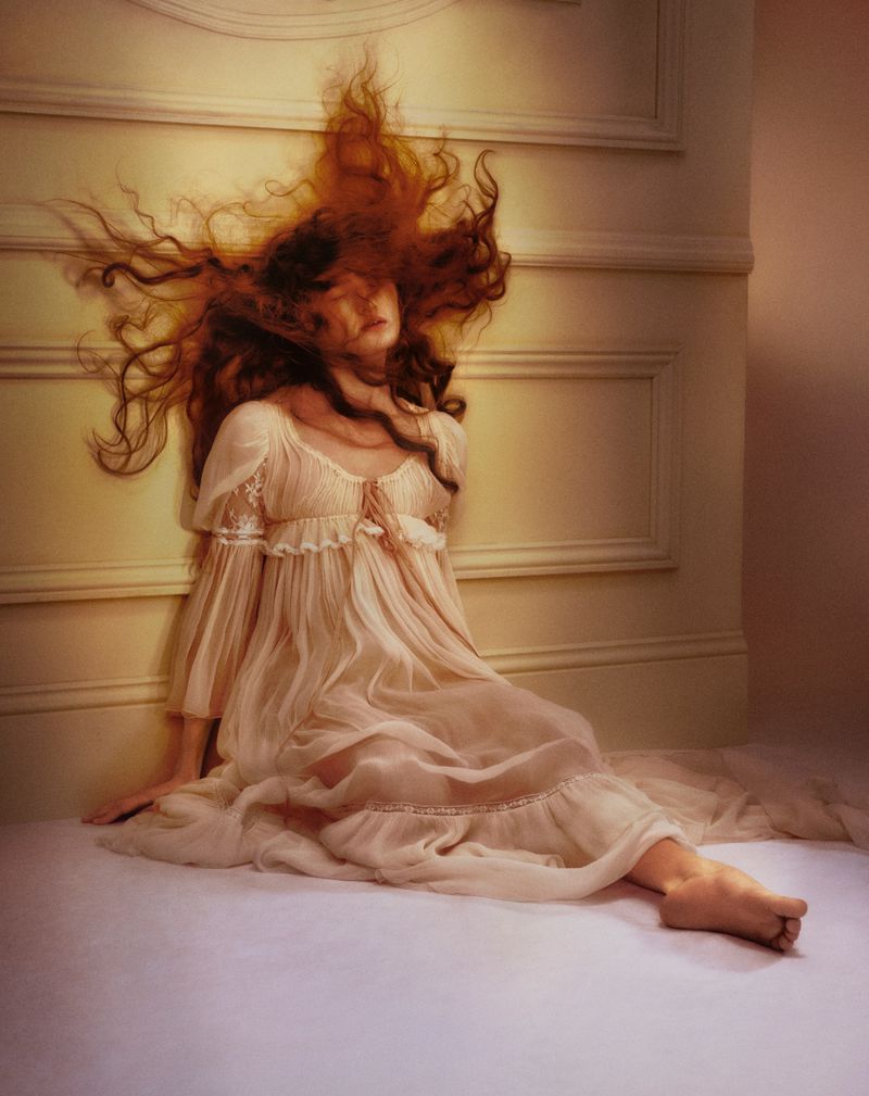 A woman with long red hair sits in a pale dress against a cream-colored wall, with a soft, dreamy aesthetic achieved through painting or effects.