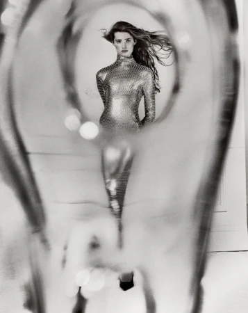 Black and white fashion shot of a woman in a metallic dress, seen through a blurred, glass-like abstract foreground.