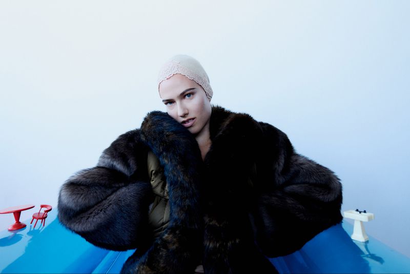 Fashion shot of a woman wearing a fur coat and beanie hat, posing on a blue surface, with miniature table and chair in the background.
