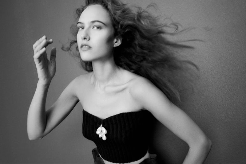 Monochrome portrait of a woman in a knitted top with a pendant, glancing upward, with windswept wavy hair against a grey background.