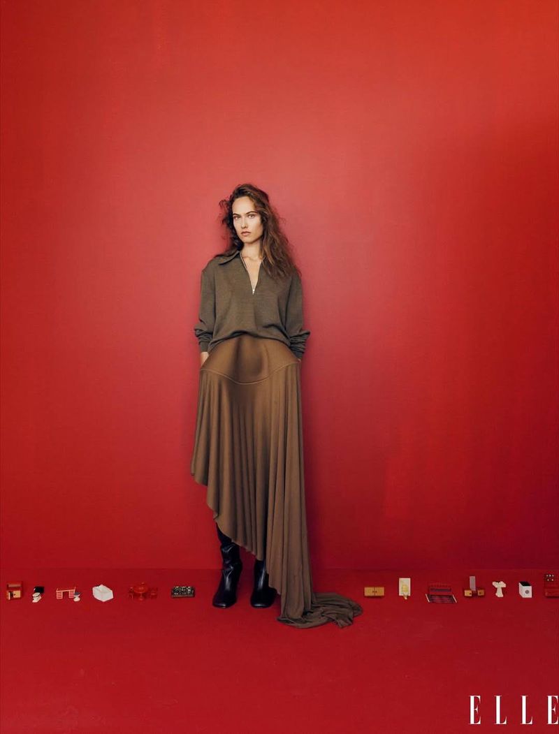 A stylish woman in an olive top and long brown skirt poses confidently against a red backdrop. She wears black boots, with 'ELLE' logo visible.