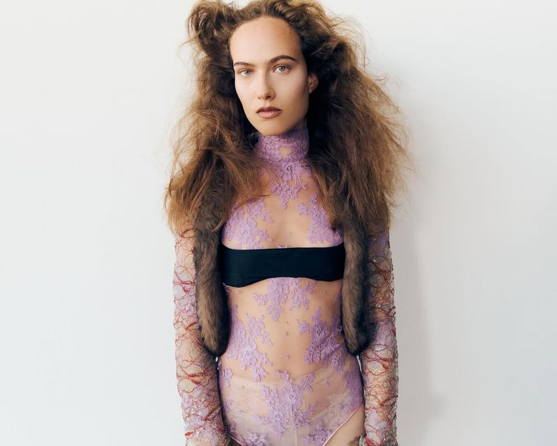 A woman with curly hair poses in a lace bodysuit with a black bandeau, against a white background. Beauty and fashion photography.
