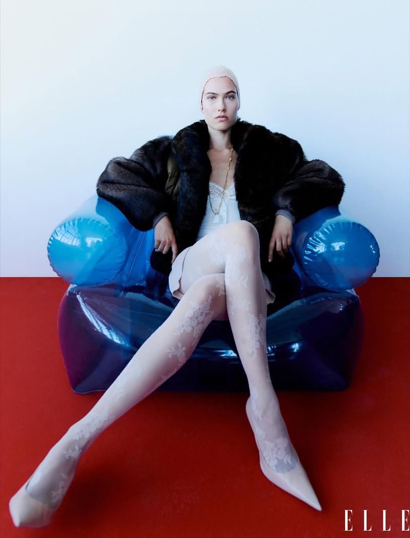 Fashion editorial image features a woman with fair skin, wearing a dark fur coat, light-pink dress, and patterned tights, sitting in a blue inflatable chair.