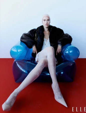 Fashion editorial image features a woman with fair skin, wearing a dark fur coat, light-pink dress, and patterned tights, sitting in a blue inflatable chair.