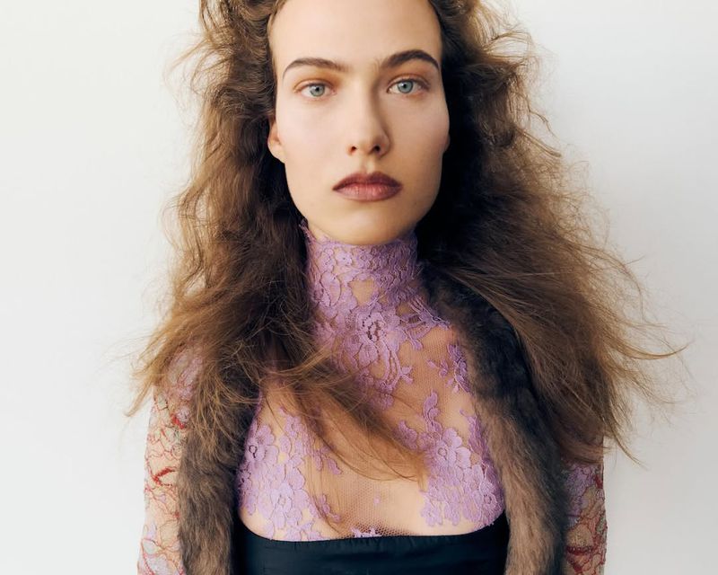 Close-up portrait of a female model with long, wavy brown hair and a sheer lilac top with floral lace detailing against a plain white backdrop.