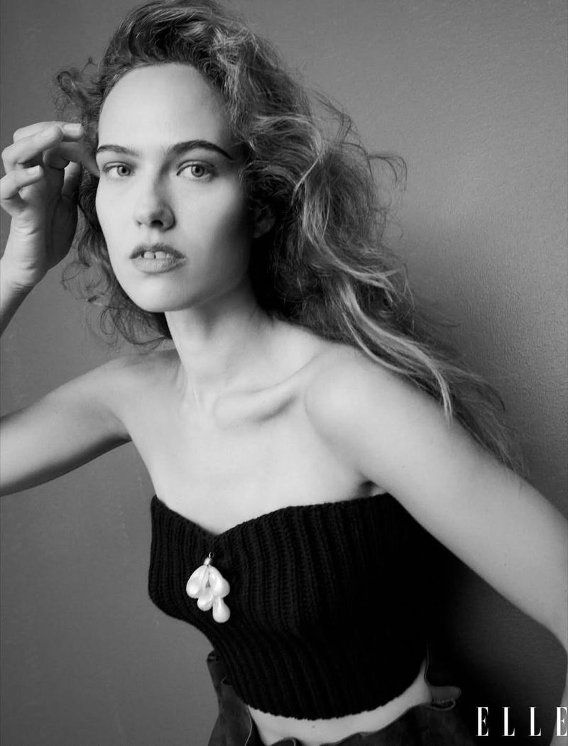 Monochrome portrait of a young woman with wavy hair, wearing a black strapless top and pearl pendant, against a neutral gray backdrop.