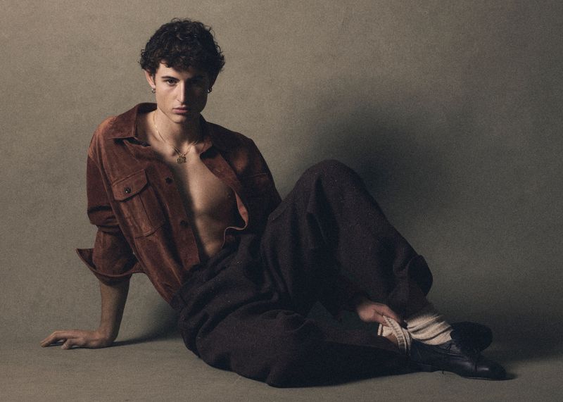 Portrait of a young man with curly hair, wearing a brown suede shirt and dark pants, seated in a relaxed pose against a muted background.
