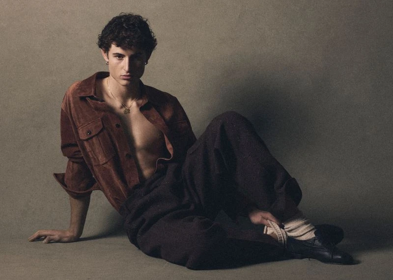 Portrait of a young man with curly hair, wearing a brown suede shirt and dark pants, seated in a relaxed pose against a muted background.