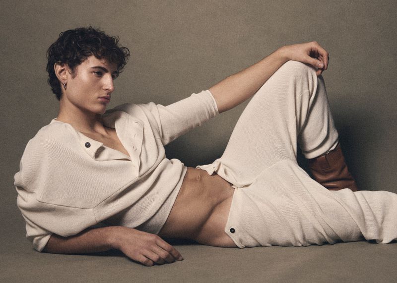 A man in a cream henley shirt and matching trousers reclining against a textured grey background for Behind the Blinds.