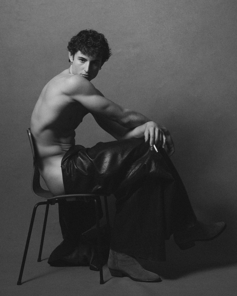 Monochrome portrait of a shirtless man seated backwards on a chair wearing dark trousers against a plain studio background.