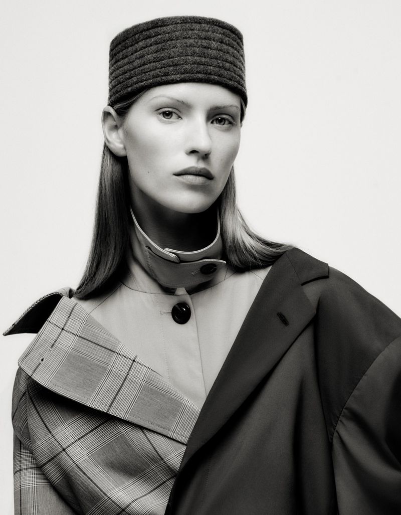 Monochrome studio portrait of a woman with straight blonde hair, wearing a layered coat with plaid and solid sections, and a gray headwrap.