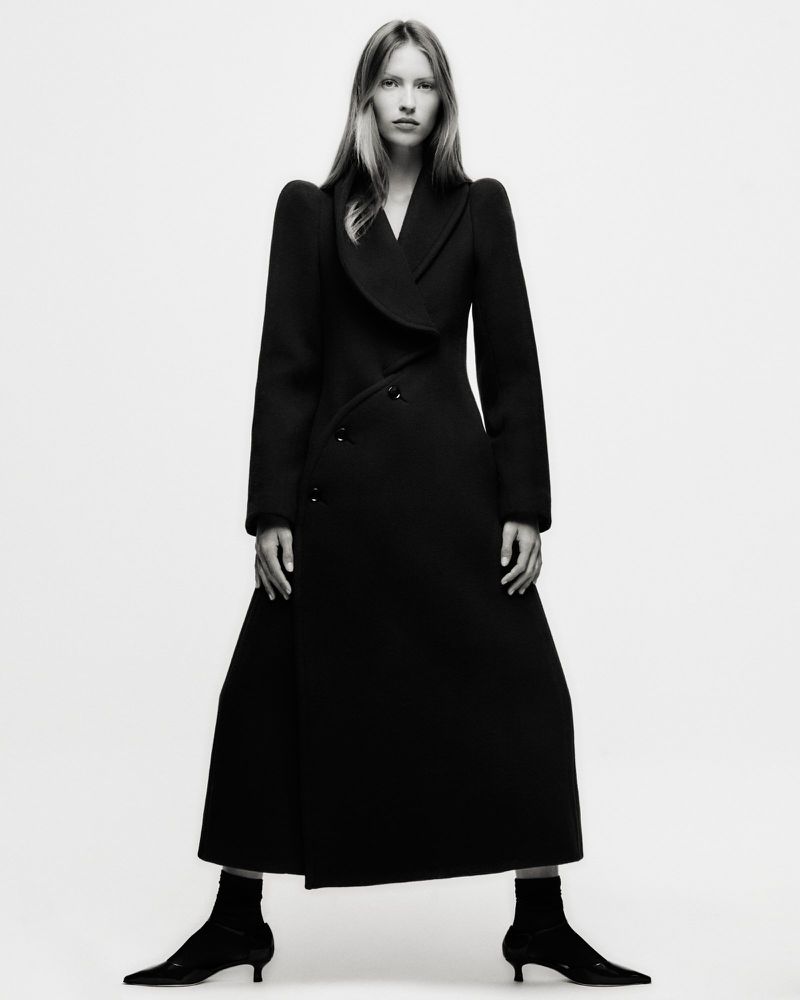 A woman in a long black coat and kitten heels stands in a photo studio. The shot is in black and white and displays a fashion editorial aesthetic.
