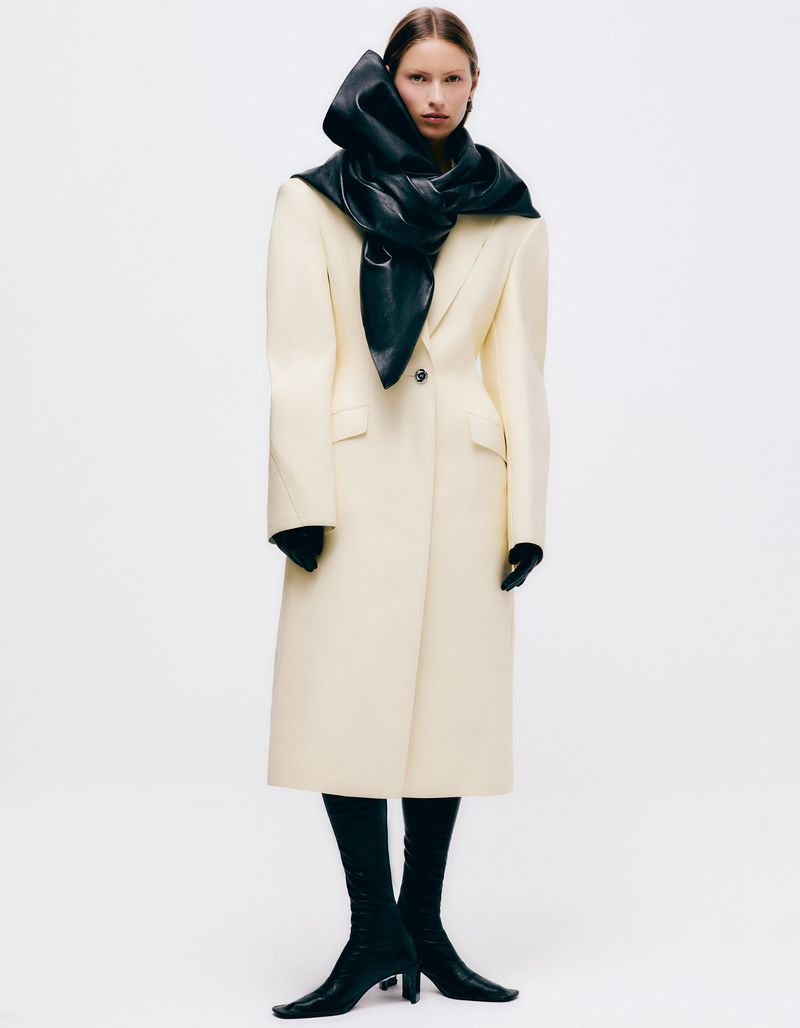 Fashion editorial shot of a woman in a long white coat, black scarf, black gloves, and boots, posing against a white backdrop.