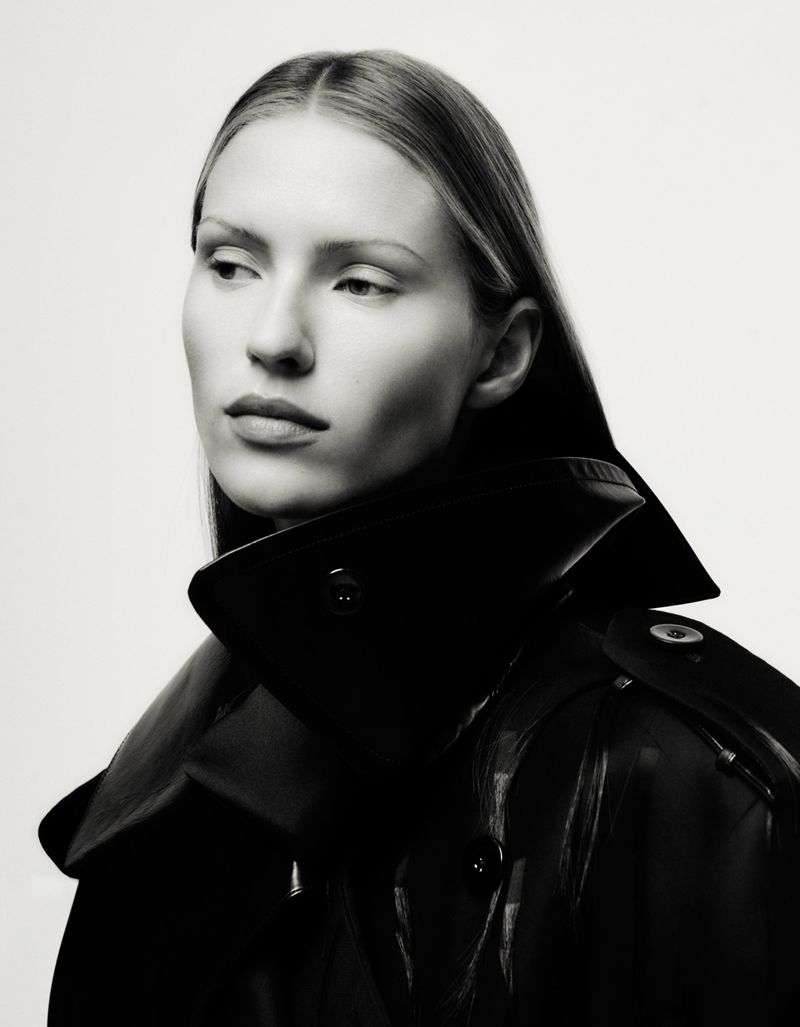 Black and white portrait of a woman in a black leather trench coat, looking off to the side against a plain white background.