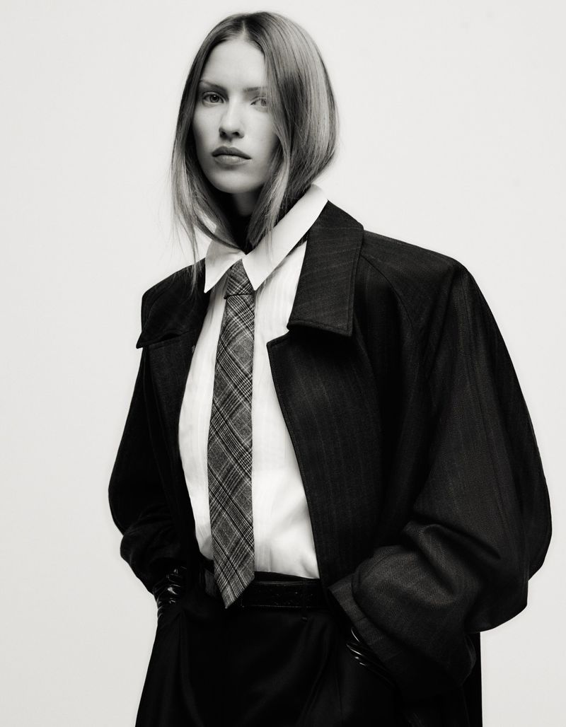 Monochrome studio portrait of a woman wearing a dark overcoat, white shirt, and plaid tie, hands in pockets against a light backdrop.