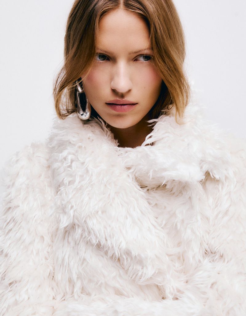 Close-up portrait of model Lea Seelenmeyer wearing a fluffy white textured coat and large silver hoop earrings against white background.