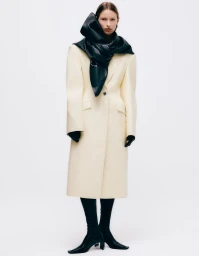Model in cream coat with large black scarf and boots against a white backdrop