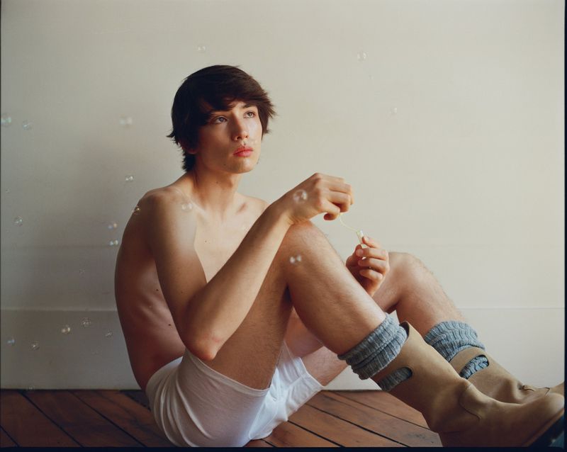 Young adult man sitting shirtless on the floor blowing bubbles inside a room with hardwood floors wearing socks and boots.