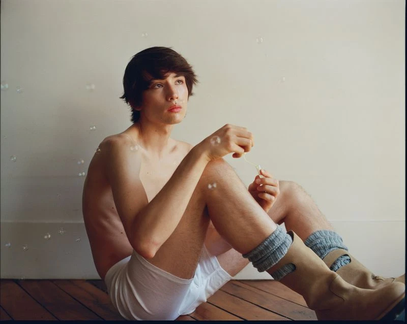 Young adult man sitting shirtless on the floor blowing bubbles inside a room with hardwood floors wearing socks and boots.