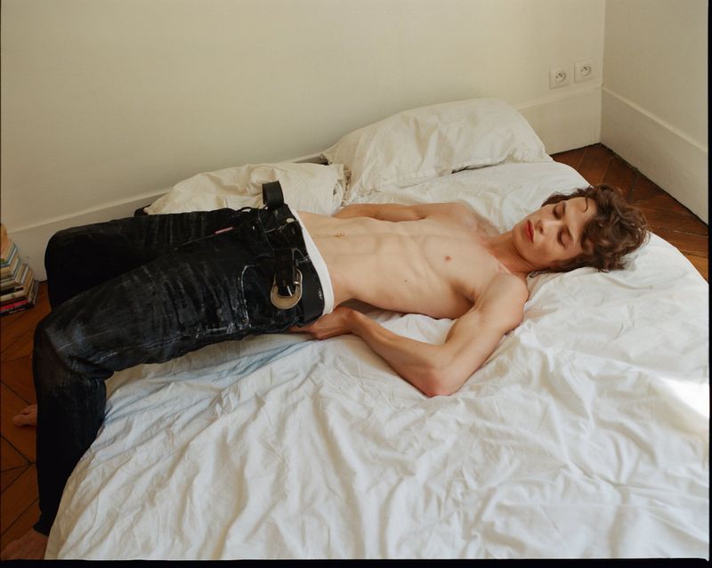 Shirtless adult male with dark hair and eyes, reclined on a white bed in jeans and a belt, in an indoor setting with bright, soft lighting.