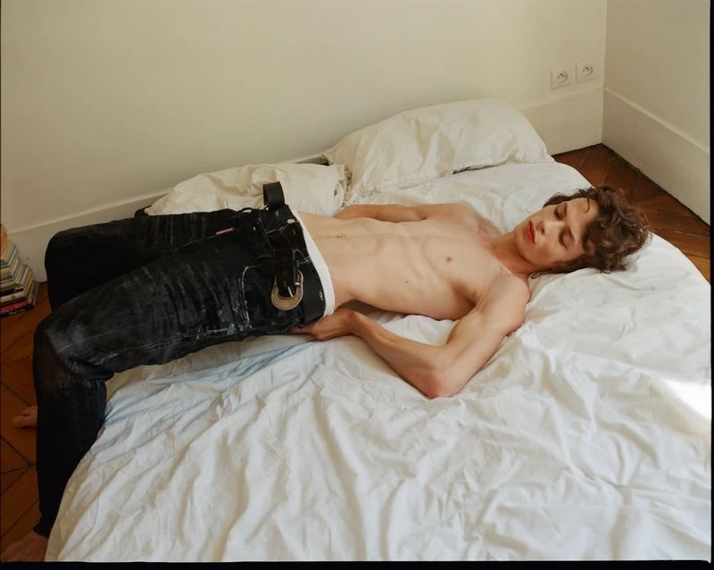 Shirtless adult male with dark hair and eyes, reclined on a white bed in jeans and a belt, in an indoor setting with bright, soft lighting.