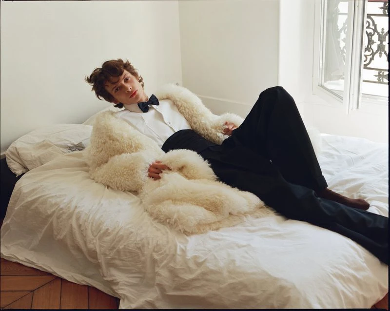A model poses in a white shirt and fur coat while reclining on a bed in a light-filled room, wearing black pants and a bow tie.
