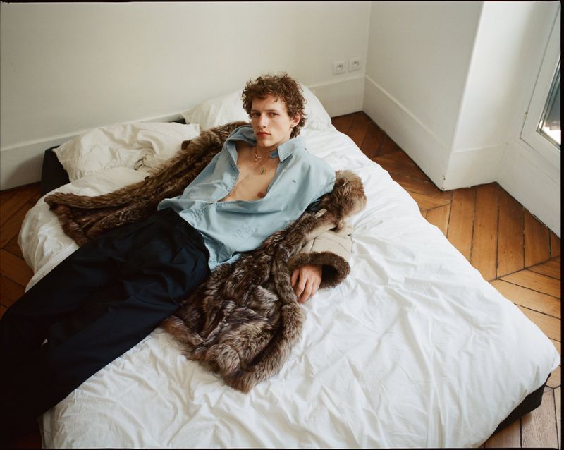 Adult male fashion model with curly hair lying on a white bed in an open shirt with a fur coat in a sunlit room.