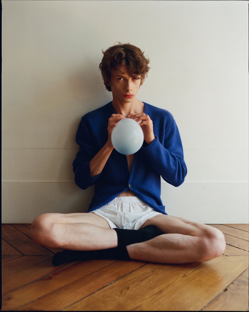 Young adult in blue cardigan sitting cross-legged on wood floor, holding light blue balloon against white wall. Indoor studio portrait.