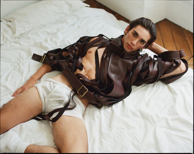 A young adult man lays on white bedsheets wearing a brown leather-like garment with straps and white underpants, a studio shot.