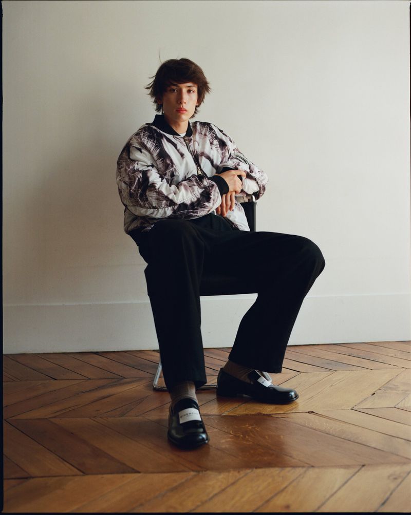 Young adult in patterned bomber jacket sits in chair on parquet floor against white wall in a fashion editorial setting.