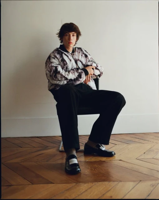 Young adult in patterned bomber jacket sits in chair on parquet floor against white wall in a fashion editorial setting.
