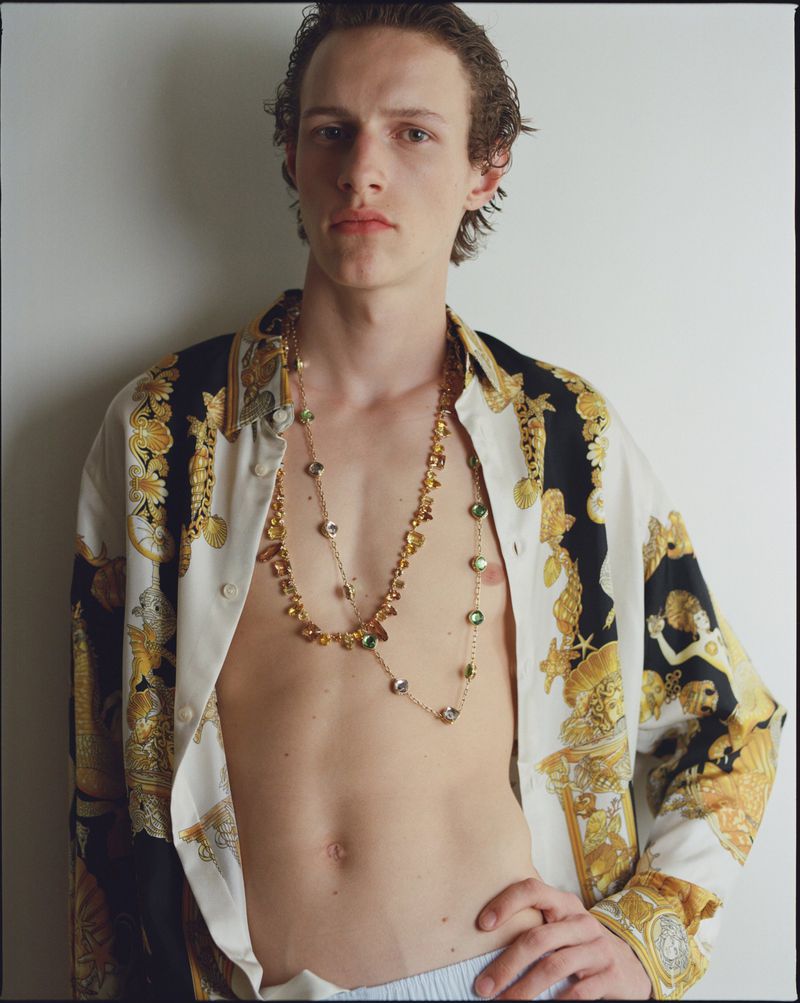 Young adult male in unbuttoned patterned shirt and jewelry poses in front of a plain wall. Fashion editorial style.