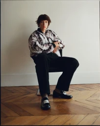 Young male model seated on a chair against a white wall wearing a patterned jacket and black trousers