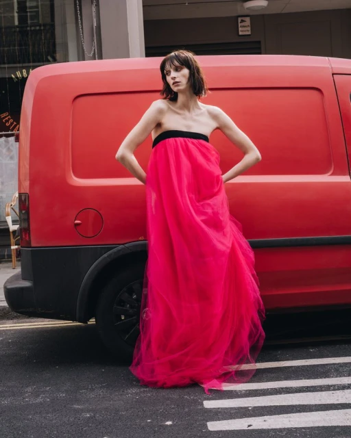 A slender woman poses in a floor-length fuchsia gown in the street, with a red van parked behind her under bright daylight.