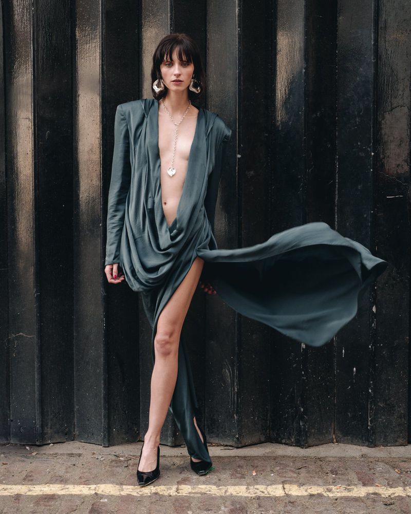 A fashionable adult female poses in a floor-length dress with an open neckline and high slit, with black high heels and large metal earrings.