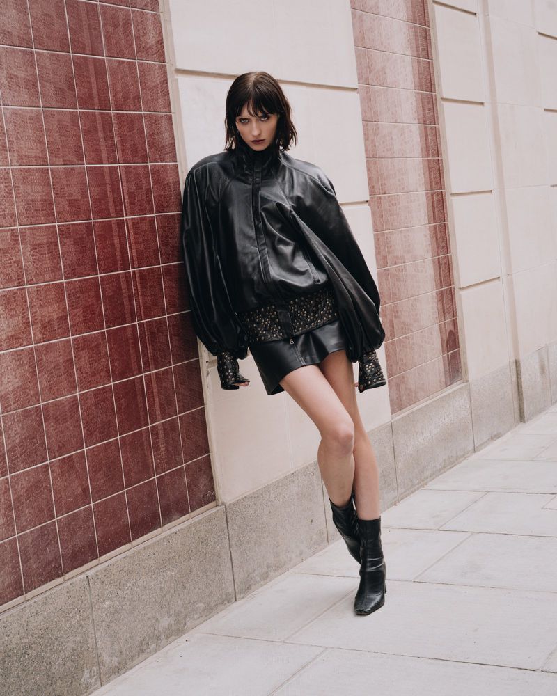 A young woman poses in a black leather jacket, short skirt and boots against a textured wall with a serious expression for a fashion shoot.