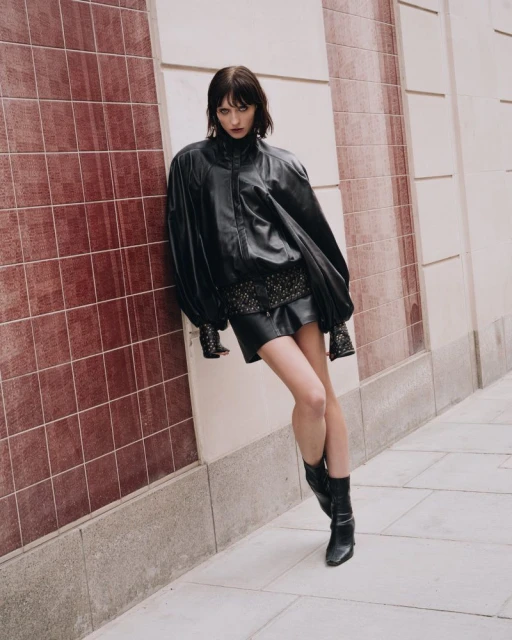 A young woman poses in a black leather jacket, short skirt and boots against a textured wall with a serious expression for a fashion shoot.