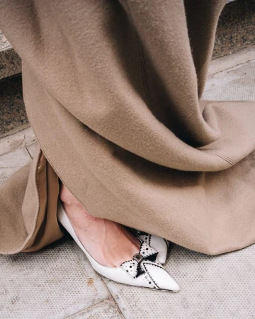 Foot in decorative white shoe with bow, tan wool coat, on grey tiled stone stairs; luxury fashion and style concept.