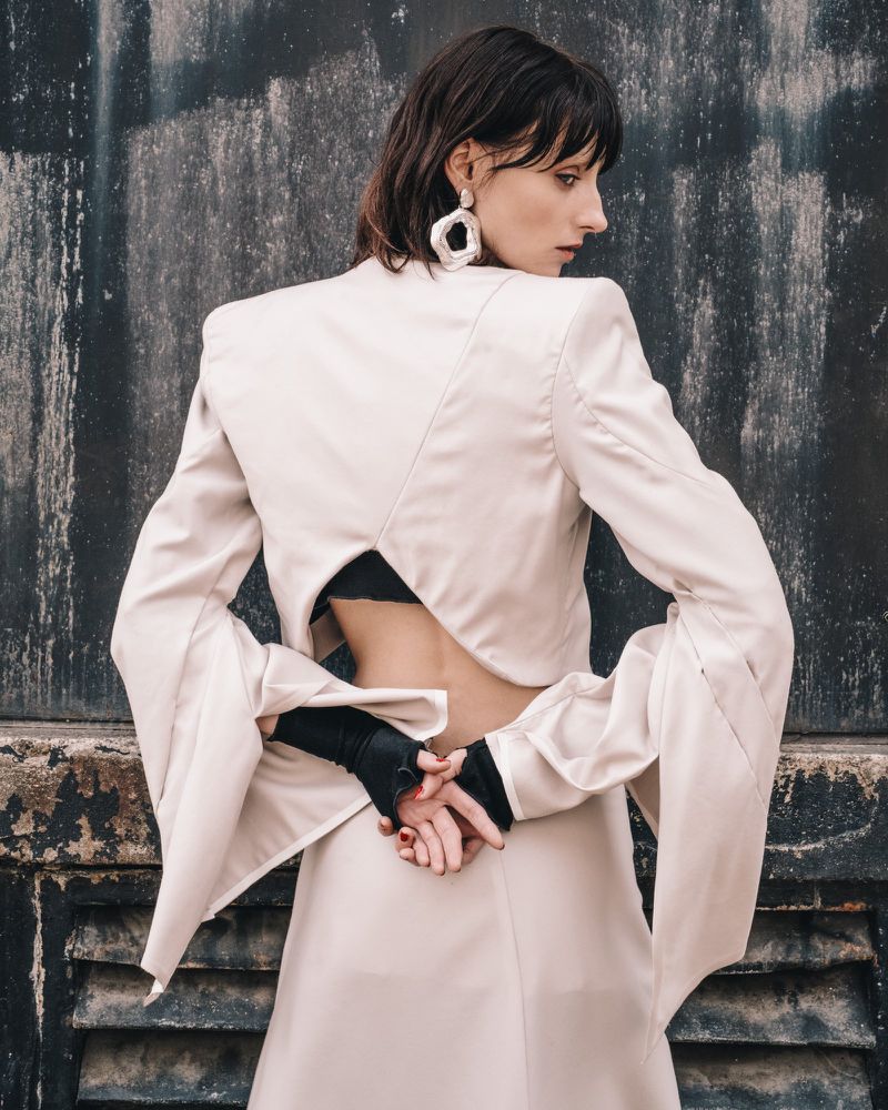 Adult female fashion model poses in a white, open-back jacket and skirt against a textured wall, showcasing modern fashion and design.