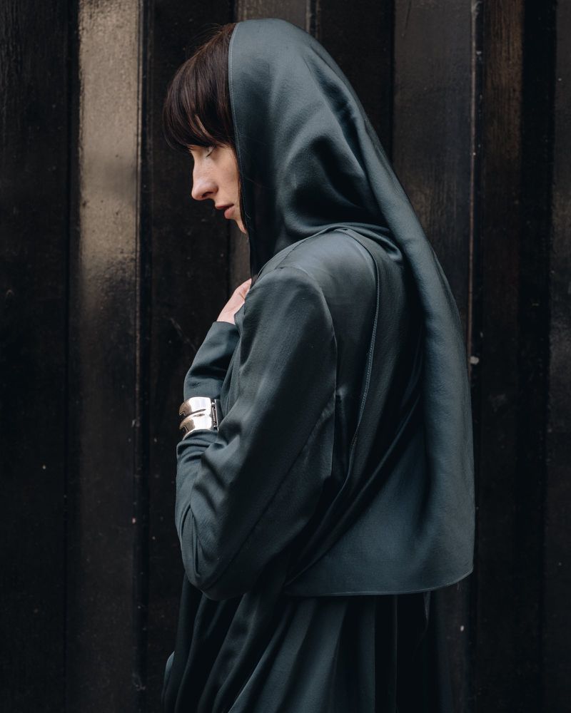 Woman in hooded teal jacket poses against dark background. Fashion portrait with soft light, textured fabric and dark tones. Profile view.