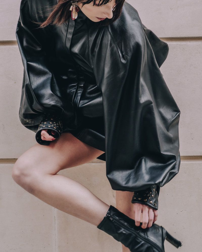 Adult female model in a black leather-like dress and black heeled boots, accessorized with earrings, poses against a neutral background.