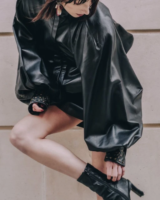 Adult female model in a black leather-like dress and black heeled boots, accessorized with earrings, poses against a neutral background.